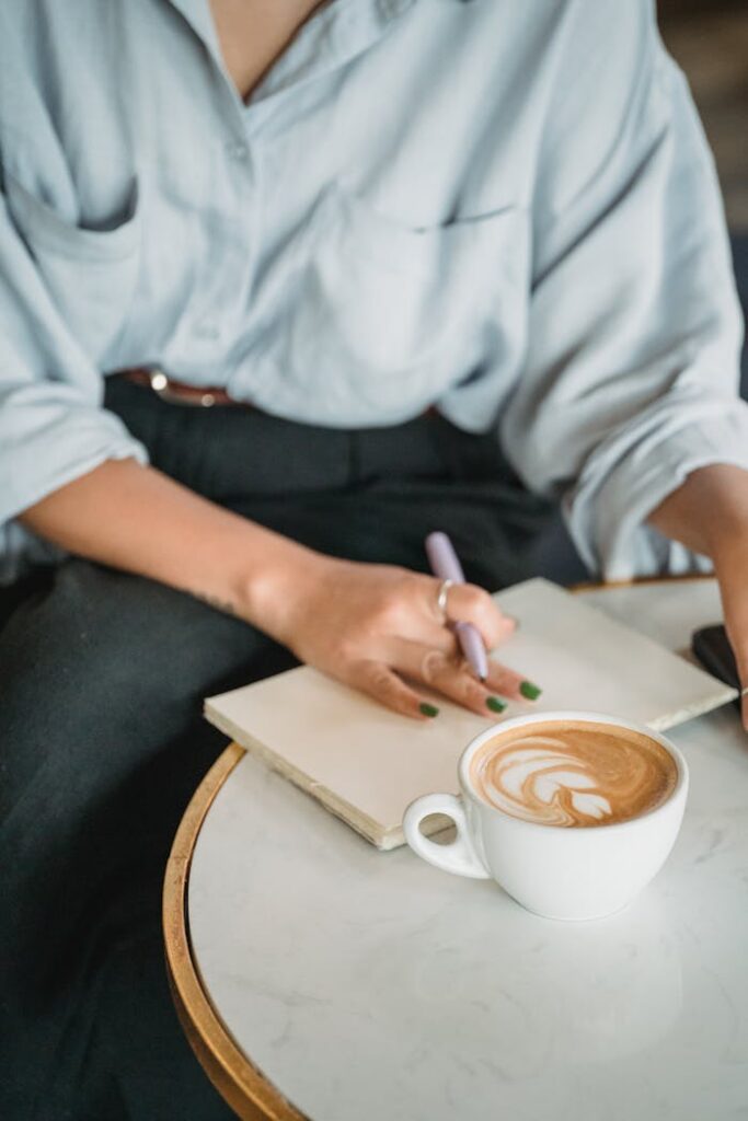pexels photo 13735945 A close-up of a woman writing in a notebook while enjoying a coffee at a café, creating a relaxed ambiance.